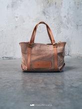 Kép betöltése a galériamegjelenítőbe: Geantă shopper maro cu bretele din piele pe podea de beton, cu perete gri pe fundal.