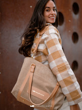 Kép betöltése a galériamegjelenítőbe: Femeie care ține o geantă bej, purtând o jachetă în carouri, pe un fundal maro texturat.