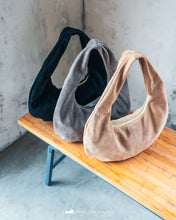 Kép betöltése a galériamegjelenítőbe: Trei genți din piele întoarsă în negru, gri și bej, așezate pe o bancă de lemn, pe fundal cu perete de beton.