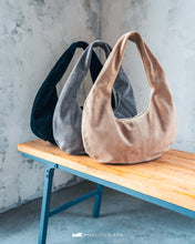 Kép betöltése a galériamegjelenítőbe: Trei genți din piele întoarsă în negru, gri și bej, așezate pe o bancă de lemn, pe fundal cu perete texturat.