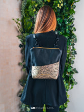 Încărcați imaginea în vizualizatorul Galeriei, Persoană care poartă un rucsac negru cu detaliu auriu decorativ, pe fundal verde cu frunziș.

